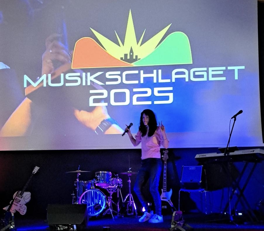 En kvinna med mikrofon står på scenen framför en skylt och instrument för evenemanget "Musikschlaget 2025".