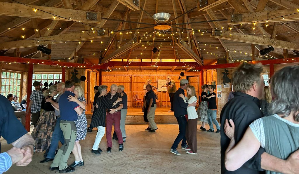 Människor dansar i par i en trähall dekorerad med ljusslingor, med musiker som spelar på scenen.