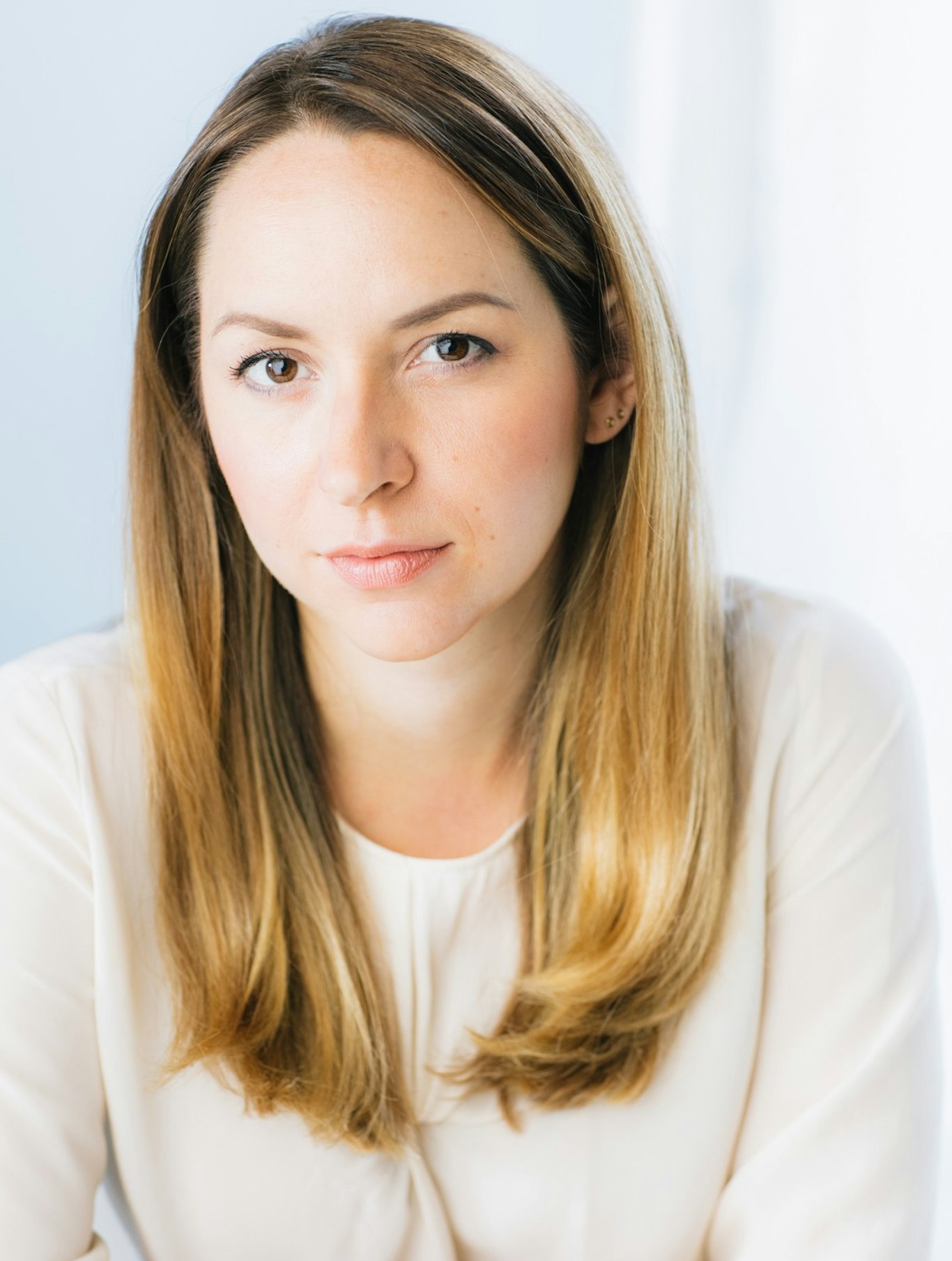 Kvinna med rakt, ljusbrunt hår i vit blus, tittar in i kameran med ett neutralt uttryck.