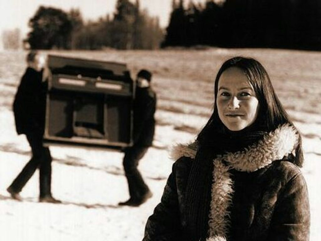 Kvinna i vinterrock ler i förgrunden, medan två personer bär ett piano över ett snöigt fält i bakgrunden.