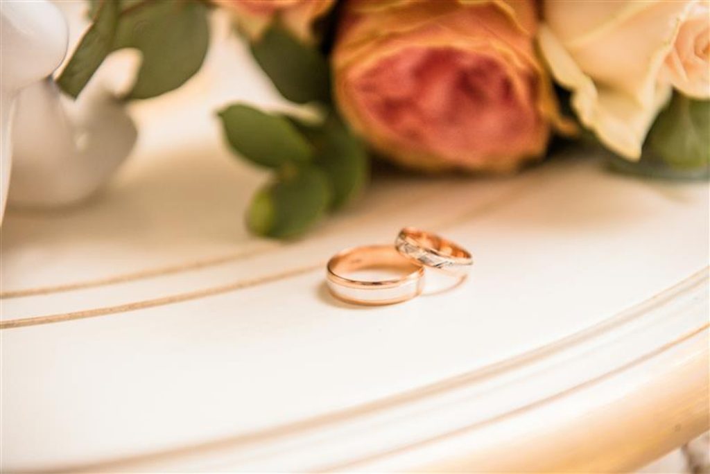 Två vigselringar i guld på en vit yta med suddiga rosor i bakgrunden.