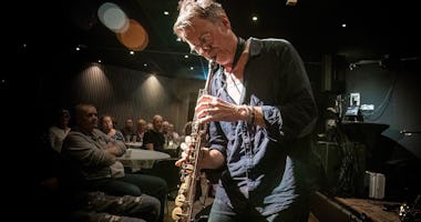 En musiker spelar saxofon på scenen framför en publik i en dunkelt upplyst lokal.