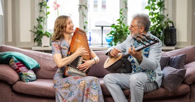 Två personer sitter i en soffa, ler och spelar stränginstrument i ett ljust, växtfyllt rum.