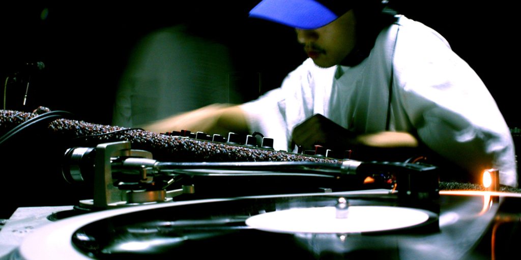 En DJ i blå keps mixar musik på skivspelare, med en vinylskiva i förgrunden.