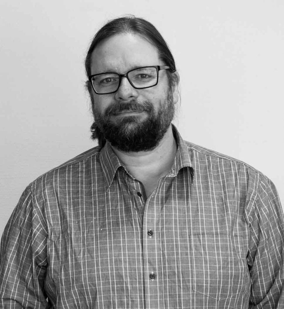 En man med glasögon och skägg, klädd i rutig skjorta, står mot en enfärgad bakgrund.
