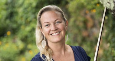 Leende kvinna med långt blont hår och vita örhängen står utomhus med grönska i bakgrunden.