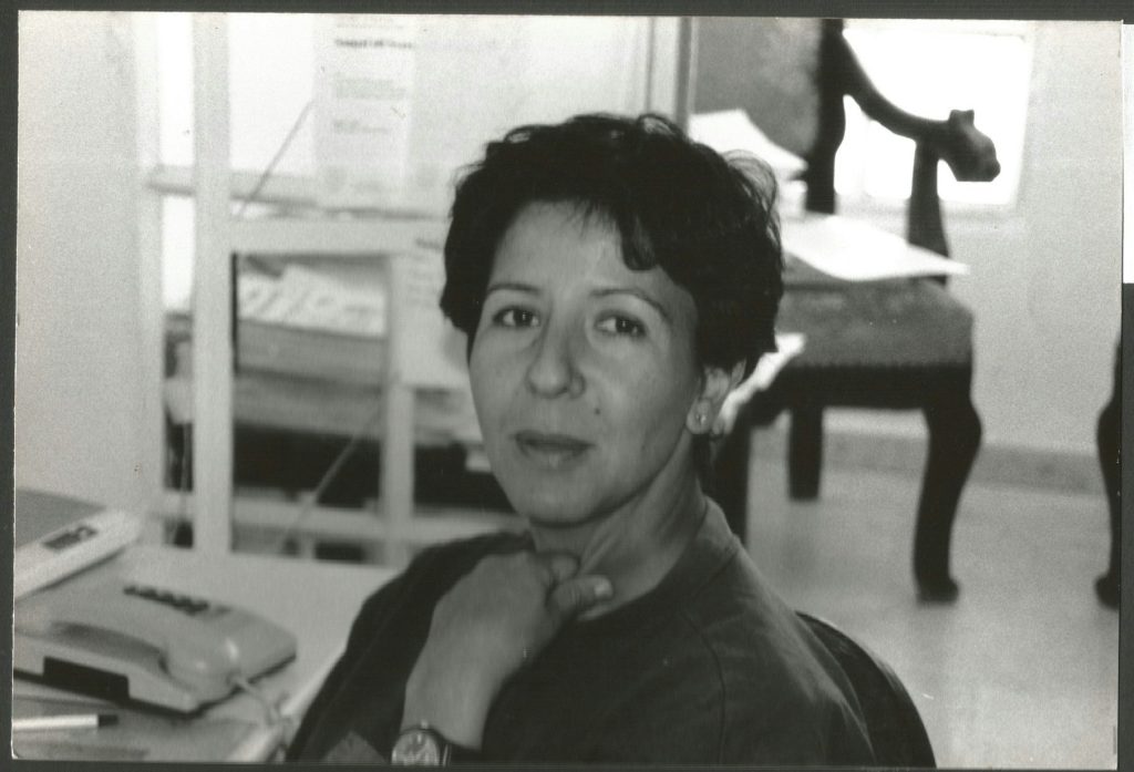 Svartvitt foto av en kvinna som sitter vid ett skrivbord och tittar in i kameran med en telefon i närheten.