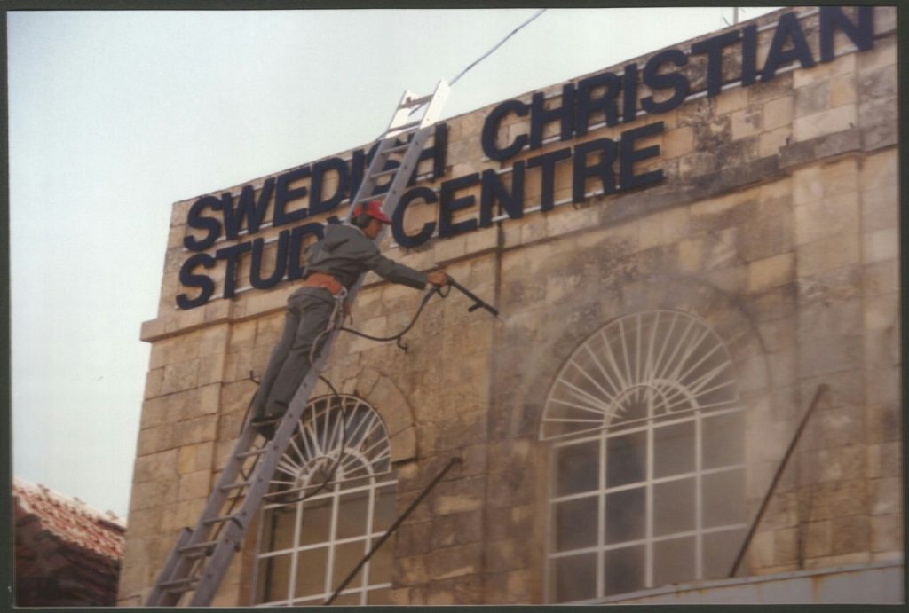 En person på en stege högtryckstvättar skylten på Sveriges kristna studiecenters byggnad.