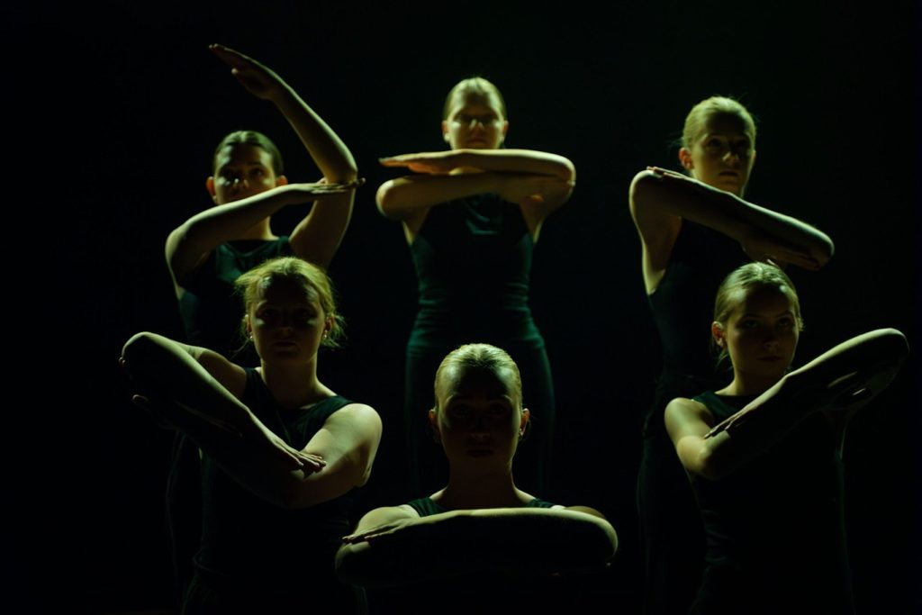 Sex dansare i mörka dräkter poserar med armarna i kors under dramatisk scenbelysning.