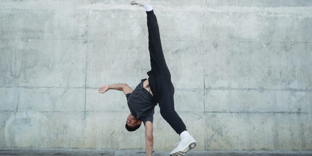 Person som utför en enhands hjulning mot en betongvägg, klädd i svarta byxor och vita skor.
