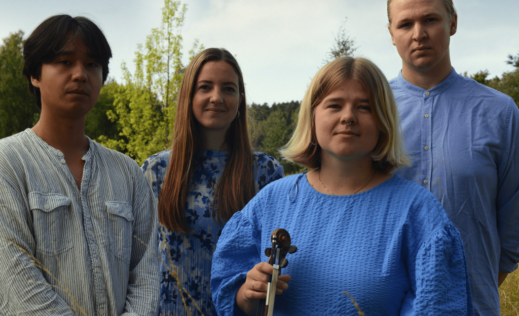 Fyra personer står utomhus i en gräsmatta, en håller i en fiol, alla bär blå eller ljusa kläder.