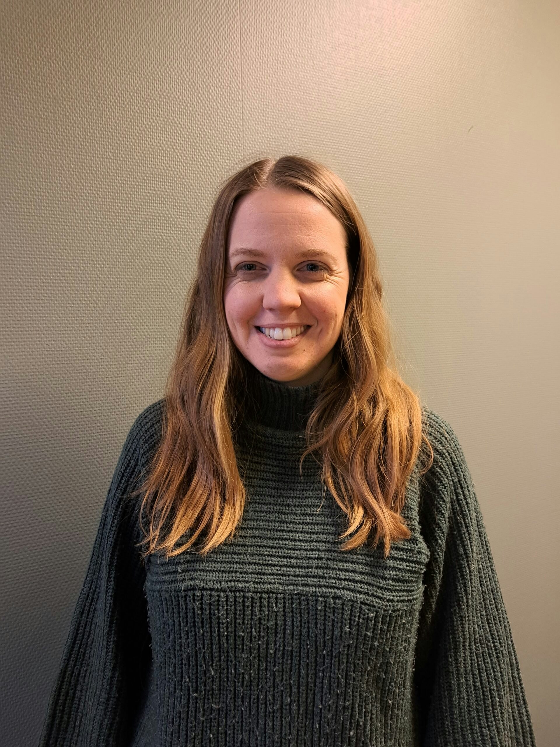 Kvinna med håret utsläppt ler in i kameran mot en grå vägg.