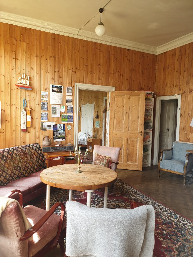 Mysigt vardagsrum med träväggar, vintagemöbler, runt bord och solljus som flödar in.