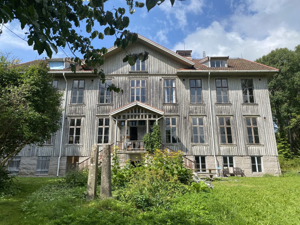 Ett stort, gammalt trähus med höga fönster och vildvuxen grönska framför, under en delvis molnig himmel.