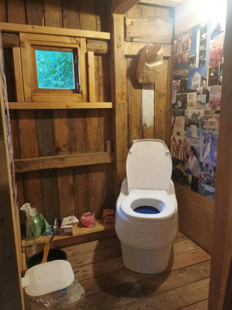 Badrum i trä med komposttoalett, litet fönster och väggar dekorerade med foton och hyllor.