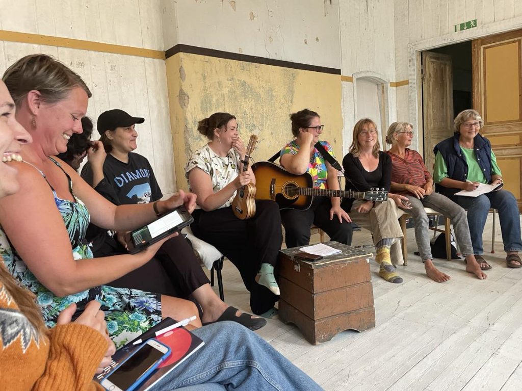 En grupp leende människor sitter inomhus, några med instrument i händerna, och sjunger och spelar musik tillsammans.