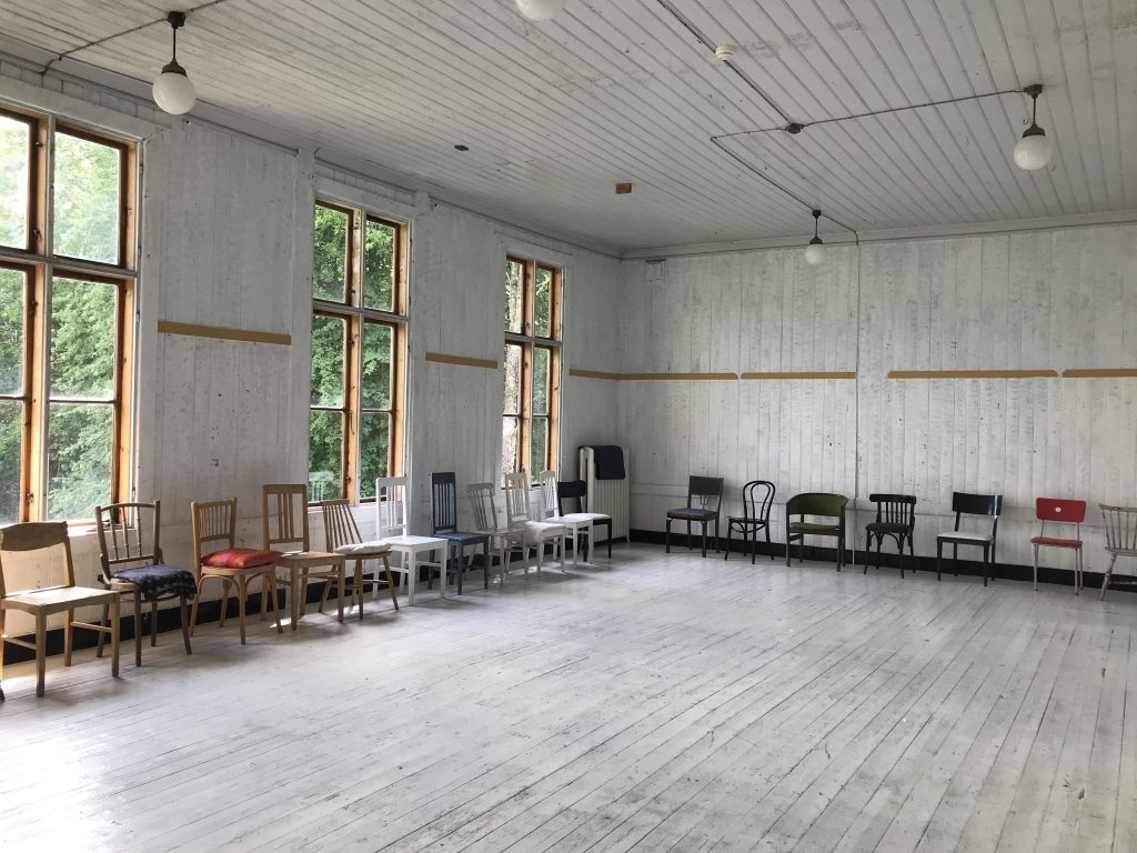 Stort, tomt rum med omaka stolar uppradade mot vita träväggar och stora fönster.