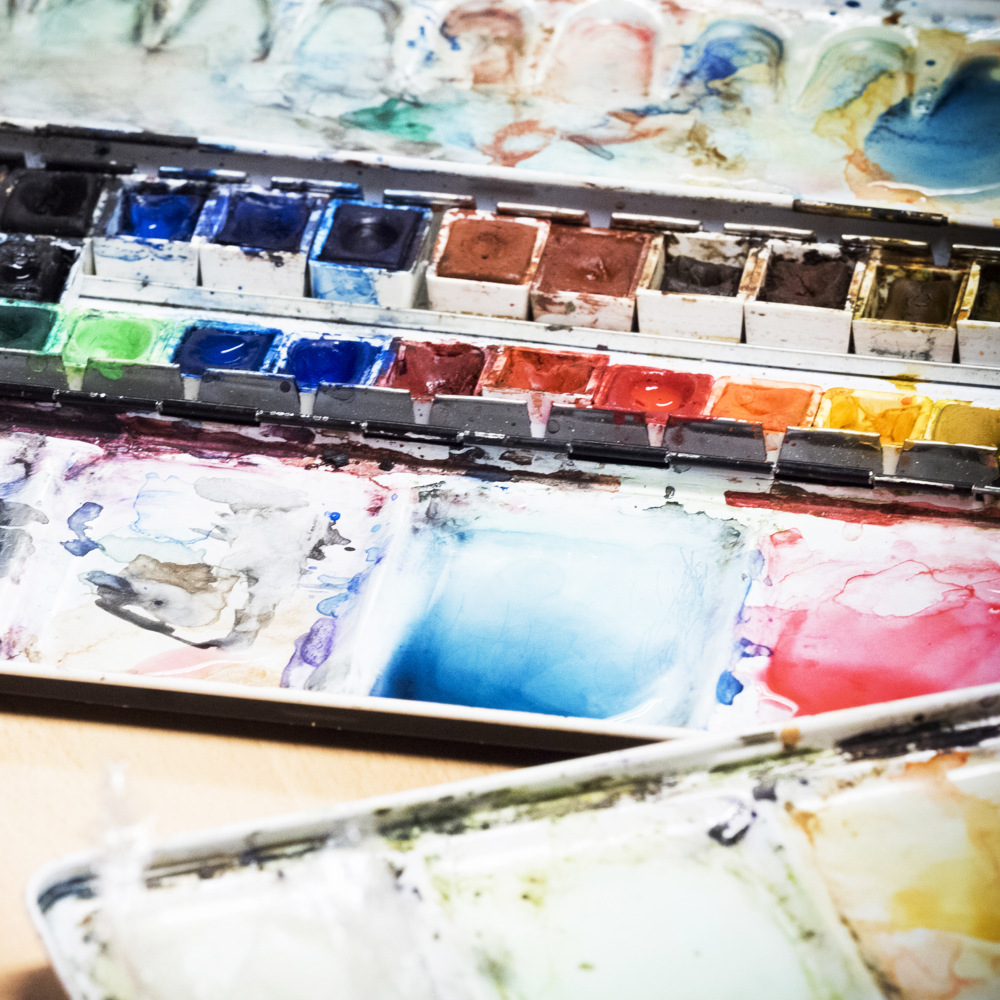 Öppen akvarellpalett med olika ljusa och blandade färger, lite färg utsmetad runt kastrullerna.