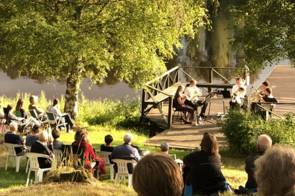 Musiker uppträder på en träbrygga vid en flod medan en publik tittar på, omgiven av träd och solljus.