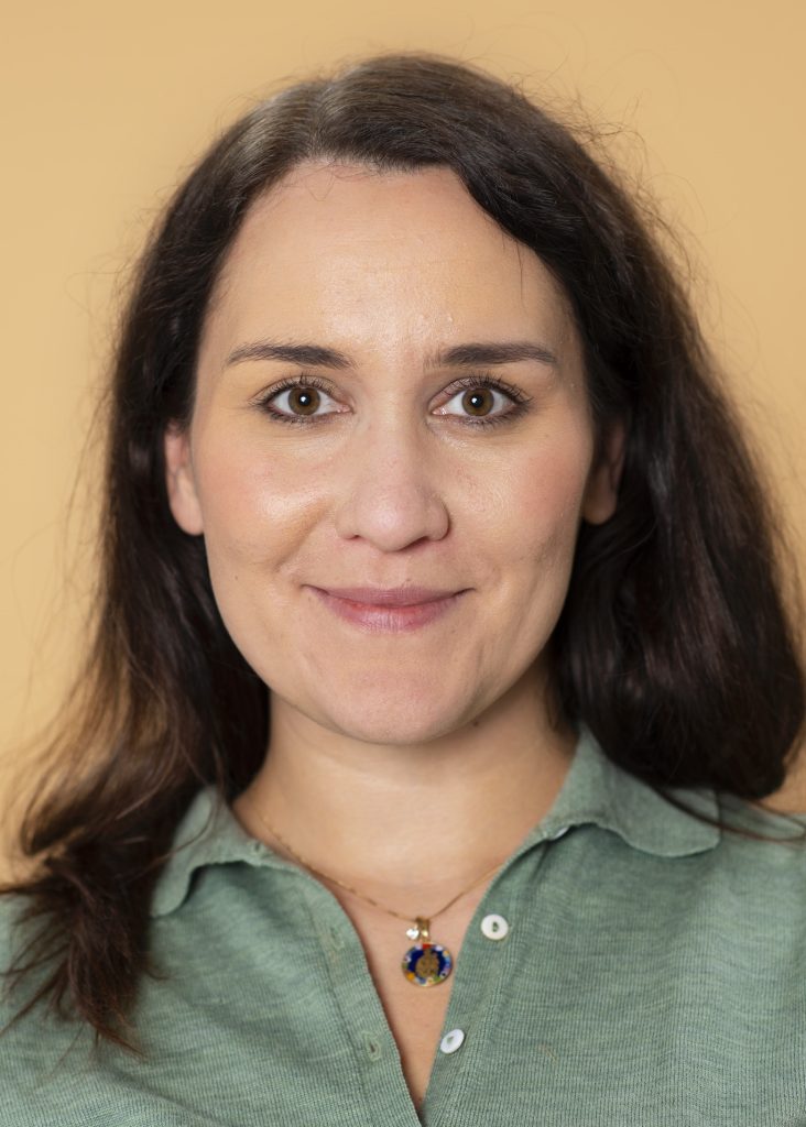 Kvinna med långt brunt hår och grön skjorta ler mot en persikofärgad bakgrund.