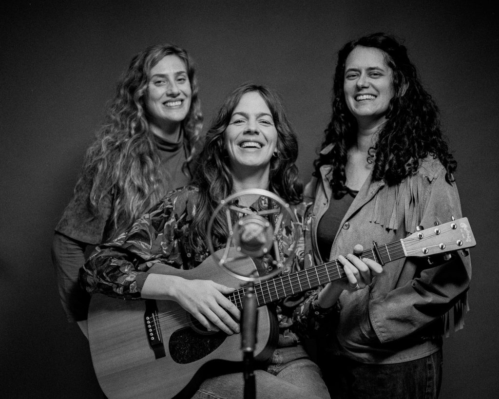 Tre leende kvinnor, varav en håller i en gitarr, poserar tillsammans framför en mikrofon mot en mörk bakgrund.