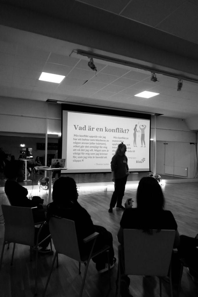 En person presenterar en bild med titeln "Vad är en konflikt?" för en publik i ett svagt upplyst rum.