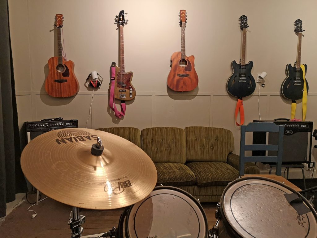 Ett trumset står framför en grön soffa med gitarrer och en bas hängande på väggen bakom.