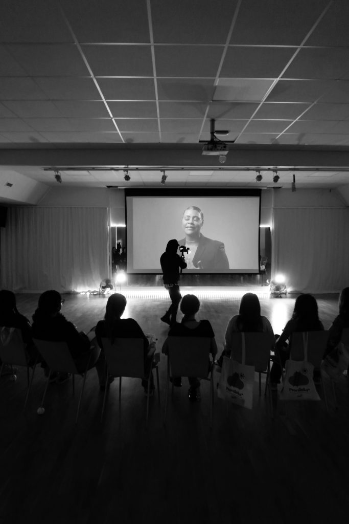 En person filmar en presentatör på en stor skärm medan en publik tittar på i ett mörkt rum.