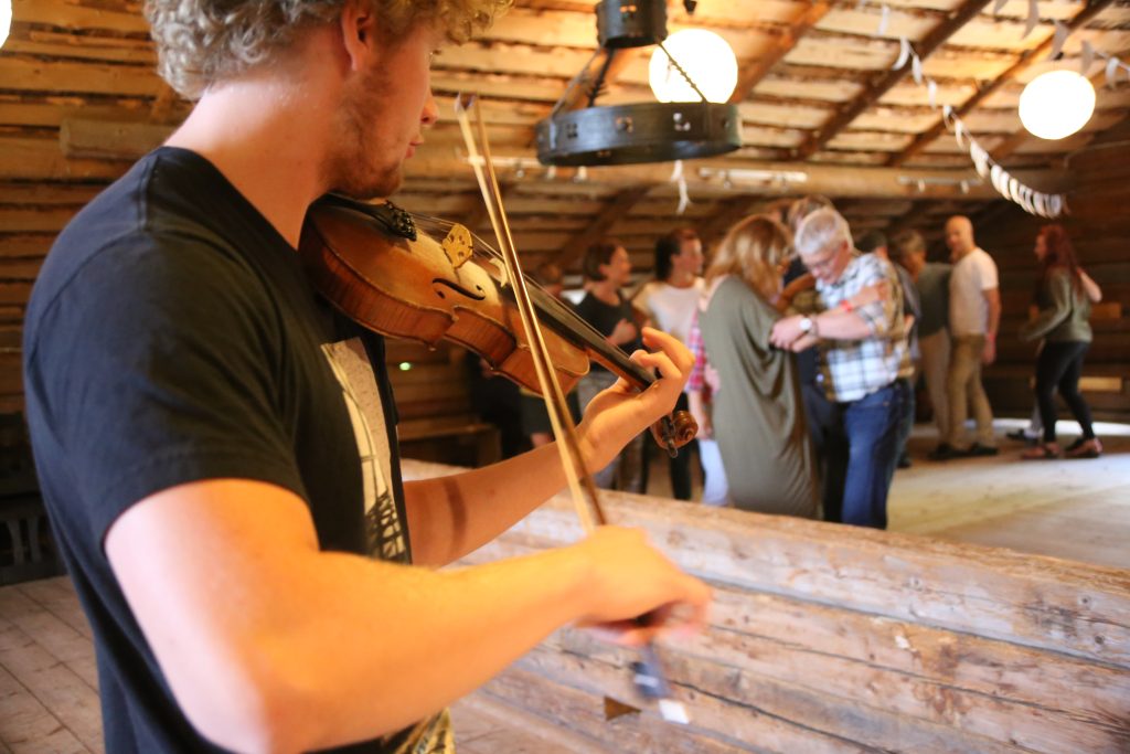 En man spelar violin i ett rum av trä medan människor dansar och umgås i bakgrunden.