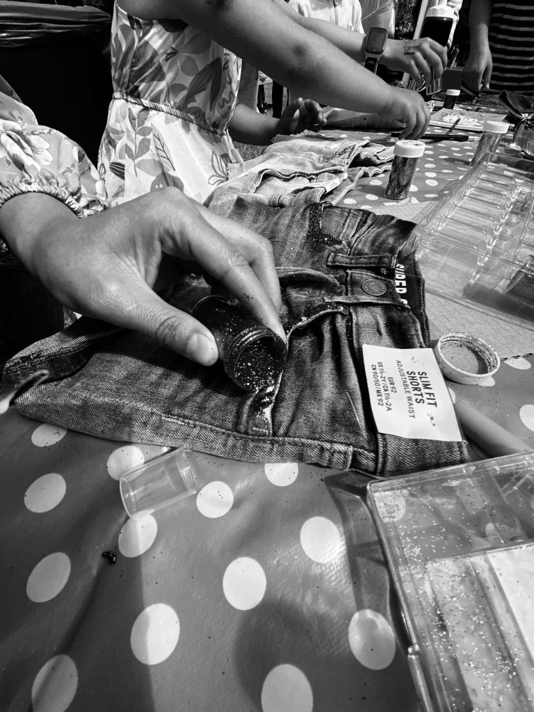 Person som dekorerar jeansshorts med glitter vid ett prickigt bord, omgiven av pysselmaterial.