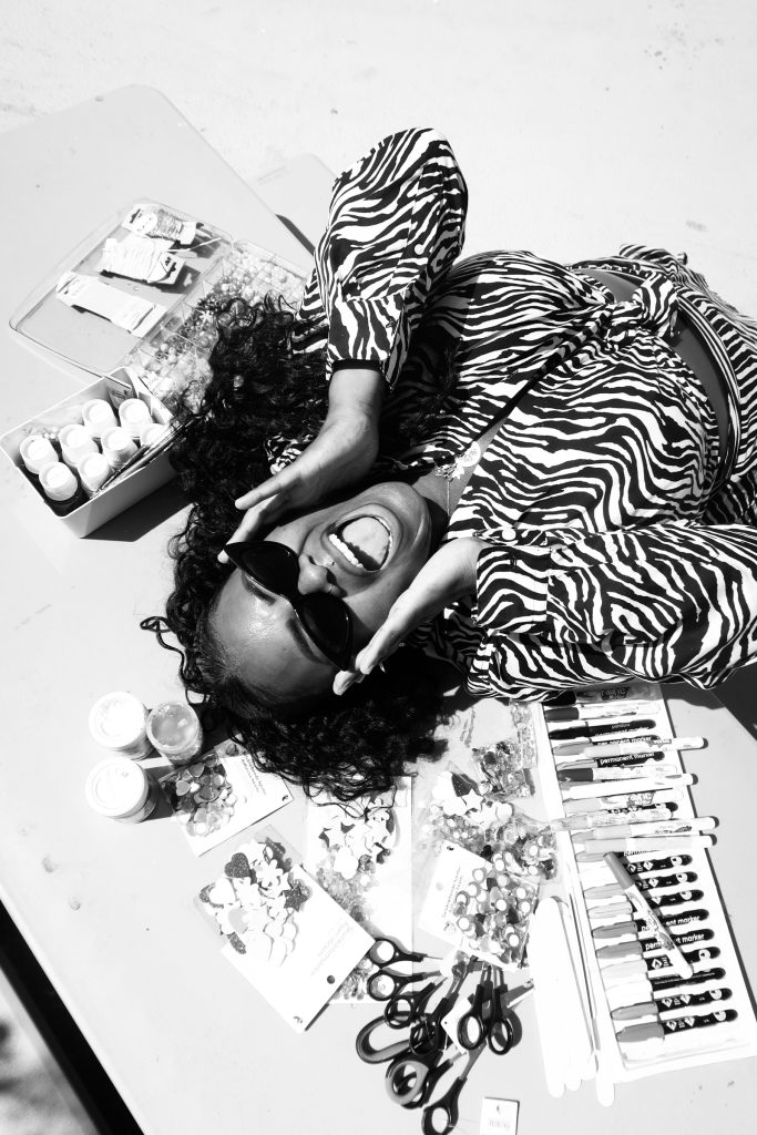 En kvinna i solglasögon ler vid ett bord med tuschpennor, målarfärg, saxar och pysselmaterial.