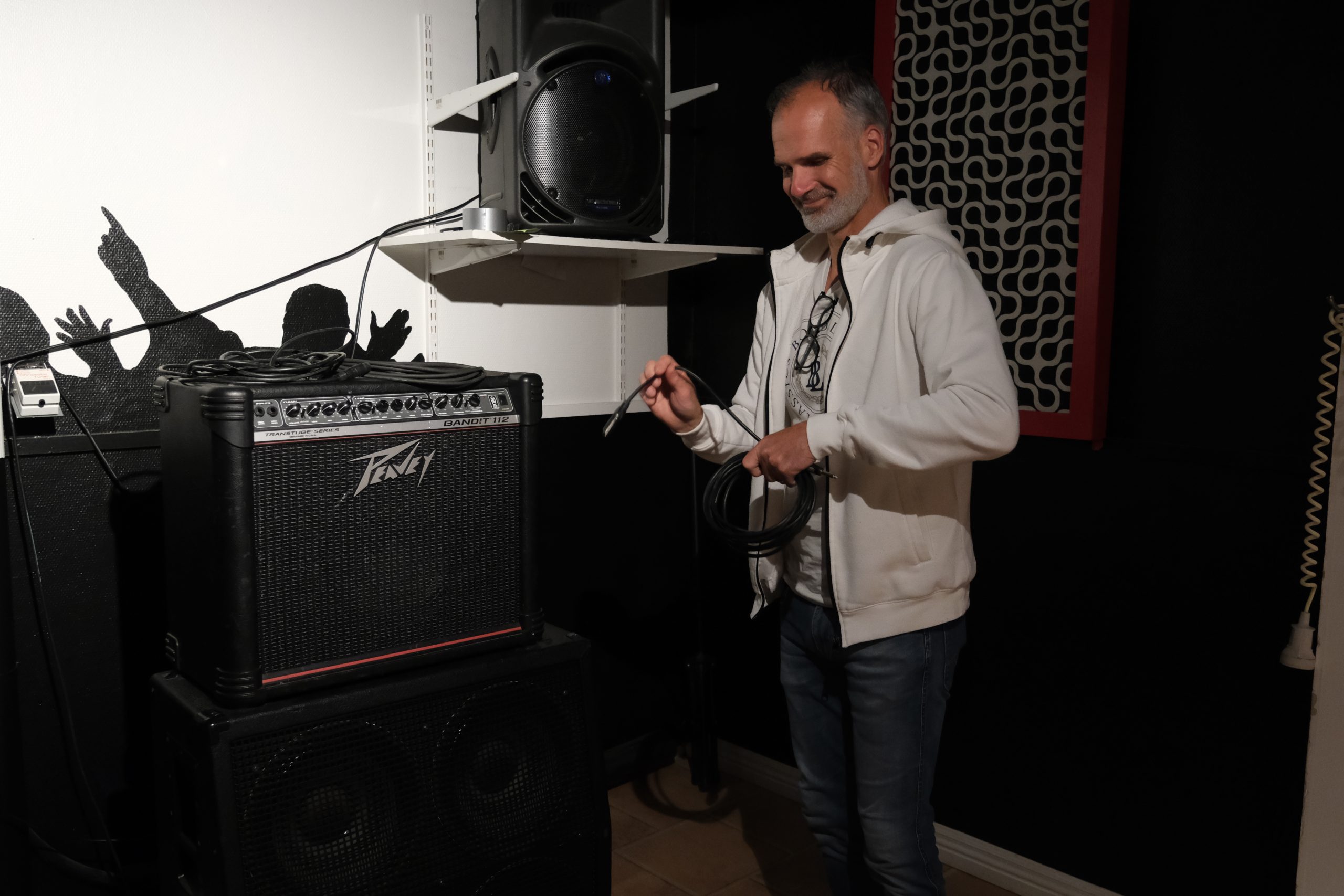 En man håller i en spiralkabel bredvid en Peavey-förstärkare i ett rum med väggkonst och högtalare.
