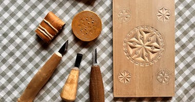 Verktyg för träsnideri, snidade stämplar i trä och en dekorerad träpanel mot en rutig tygbakgrund.