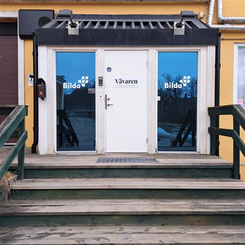 Entré till en gul byggnad med vit dubbeldörr och skyltarna "Vävaren" och "Bilda" på glaset.
