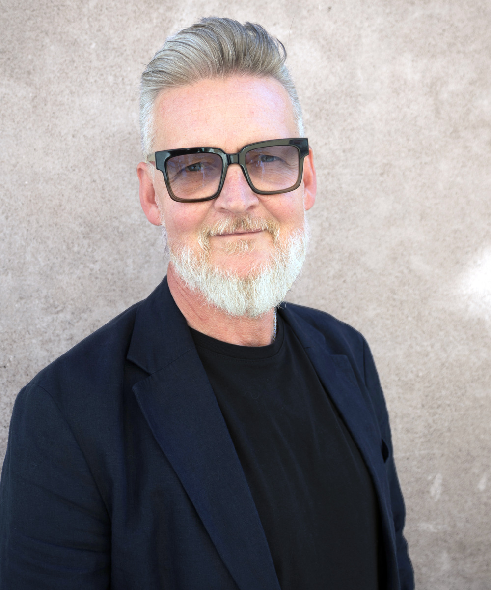 Leende man med grått hår och skägg, klädd i glasögon, svart skjorta och mörk kavaj, mot beige vägg.