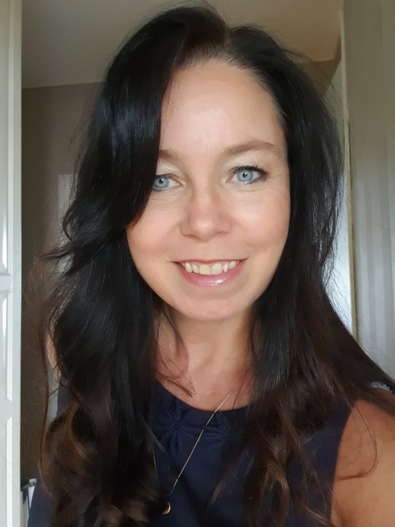 En kvinna med långt mörkt hår och blå ögon ler mot kameran i ett väl upplyst rum.