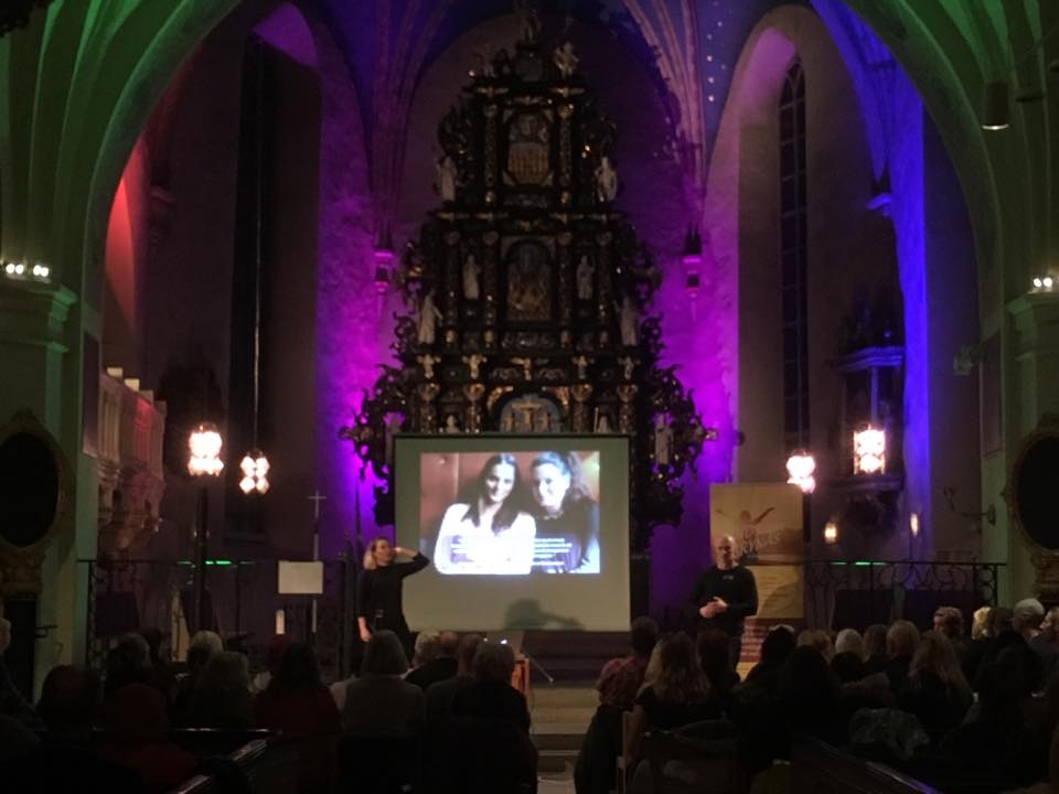 En publik tittar på en presentation i en kyrka med färgglad belysning och ett stort utsmyckat altare i bakgrunden.