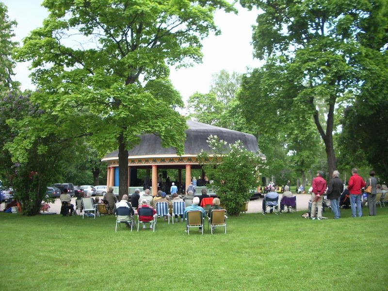 Människor sitter på stolar utomhus och tittar på en föreställning i en paviljong som omges av gröna träd och gräs.