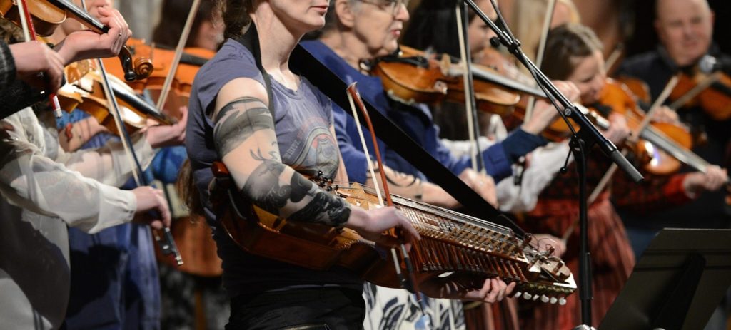 Människor spelar stränginstrument på scenen, bland annat en tatuerad kvinna längst fram som håller i en nyckelharpa.
