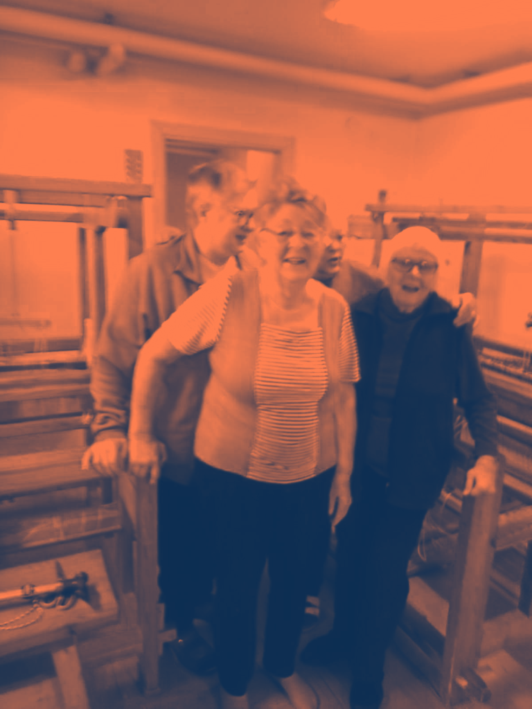 Tre leende äldre vuxna står tillsammans inomhus vid vävstolar av trä och poserar för ett foto.