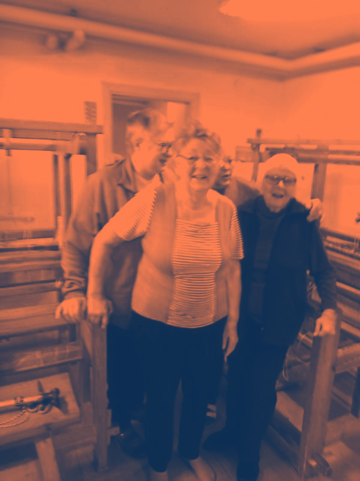 Tre leende äldre vuxna står tillsammans inomhus vid vävstolar av trä och poserar för ett foto.