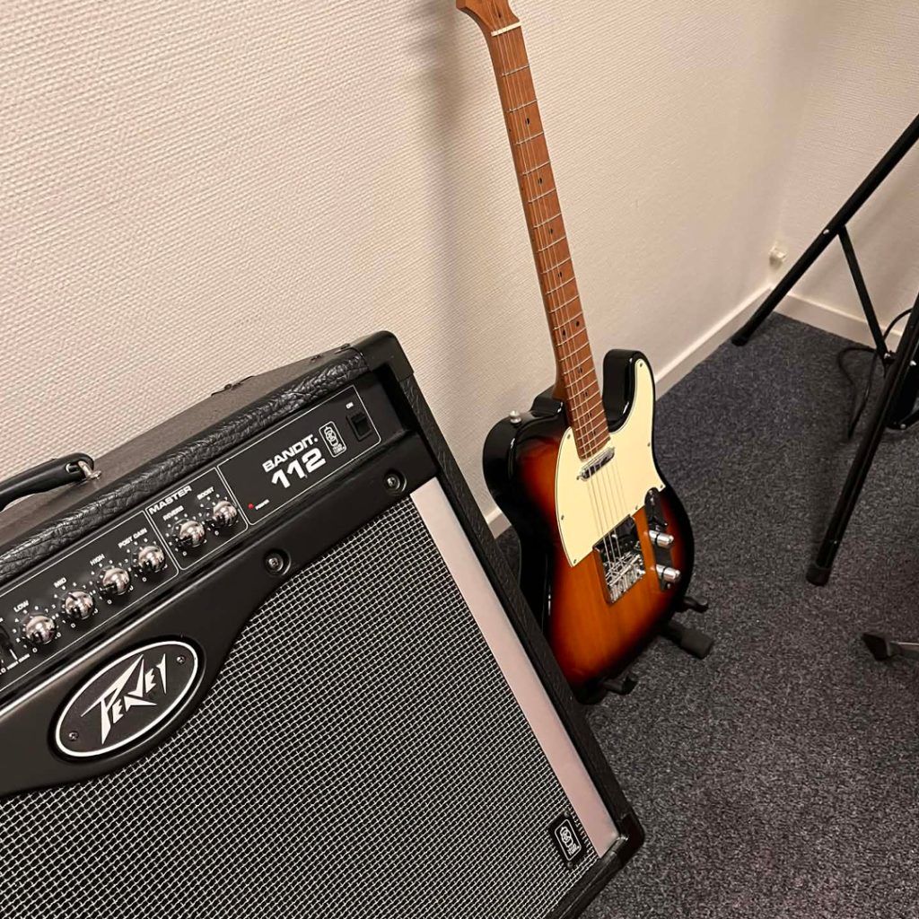 En sunburst elgitarr står lutad mot en vägg bredvid en Peavey Bandit 112-förstärkare på en golvmatta.