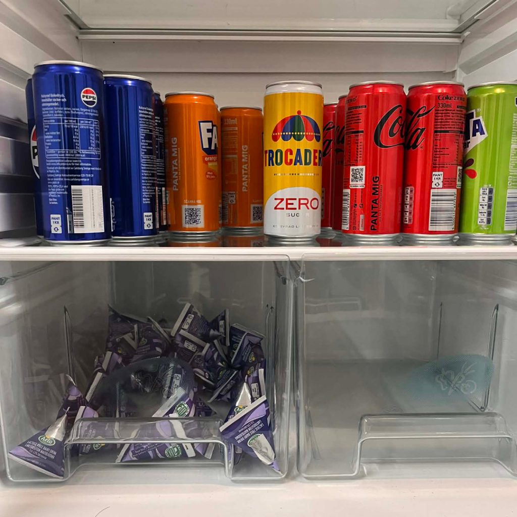 Rader av olika läskburkar på en kylskåpshylla, med små lila krämpaket i lådorna under.