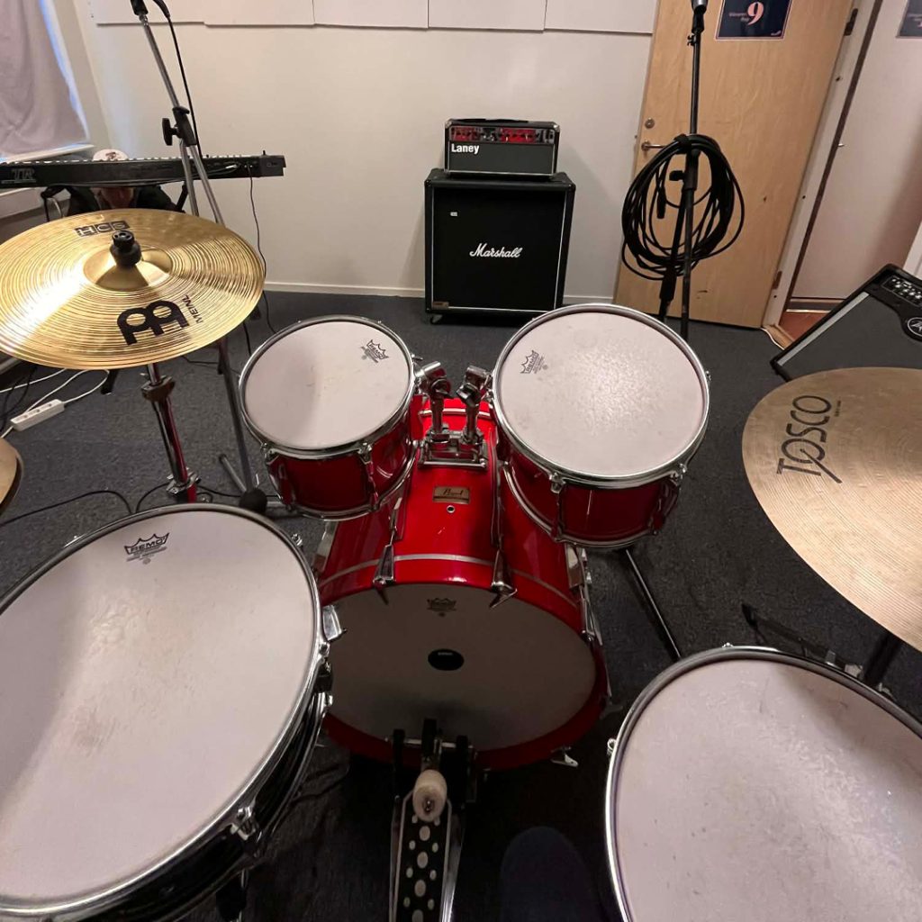 Vy bakifrån över ett rött trumset med cymbaler och förstärkare i en replokal för musik.