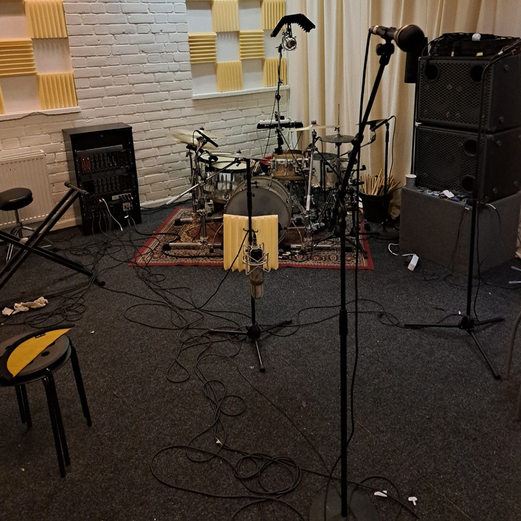 Ett musikrepetitionsrum med mikrofoner, ett trumset, högtalare och kablar utspridda på golvet.