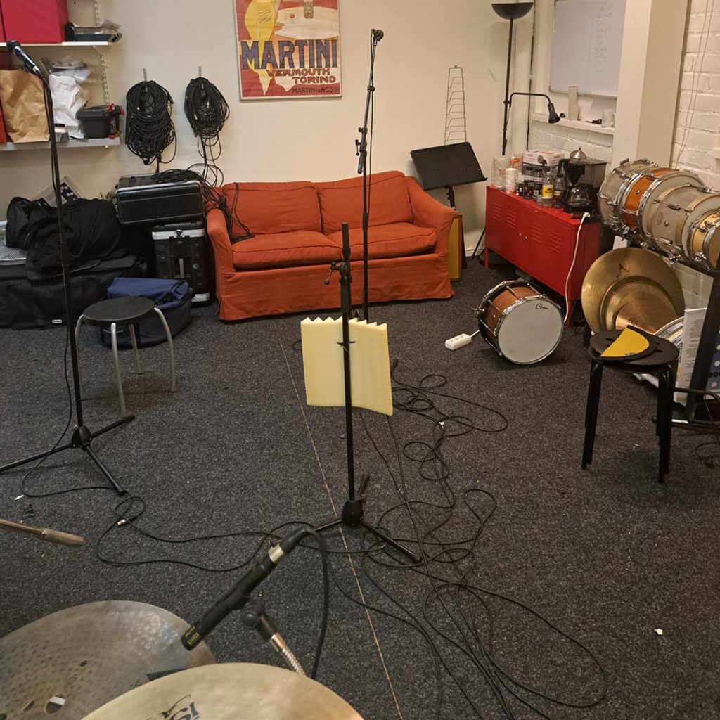 Ett musikrepetitionsrum med en röd soffa, trummor, cymbaler, mikrofoner och kablar på heltäckningsmattan.