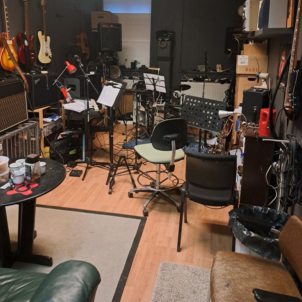 En rörig musikstudio med gitarrer, stolar, notställ och olika instrument och utrustning utspridda.