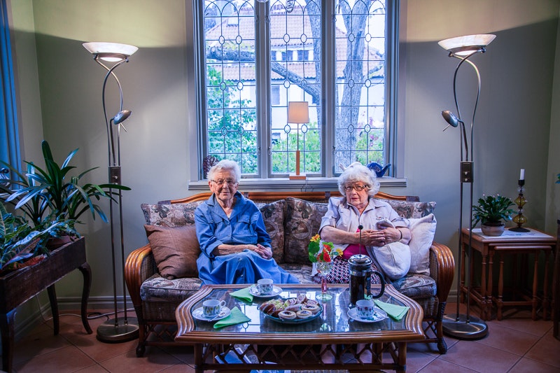Två äldre kvinnor sitter i en soffa i ett mysigt vardagsrum, med snacks och drycker på soffbordet i glas.