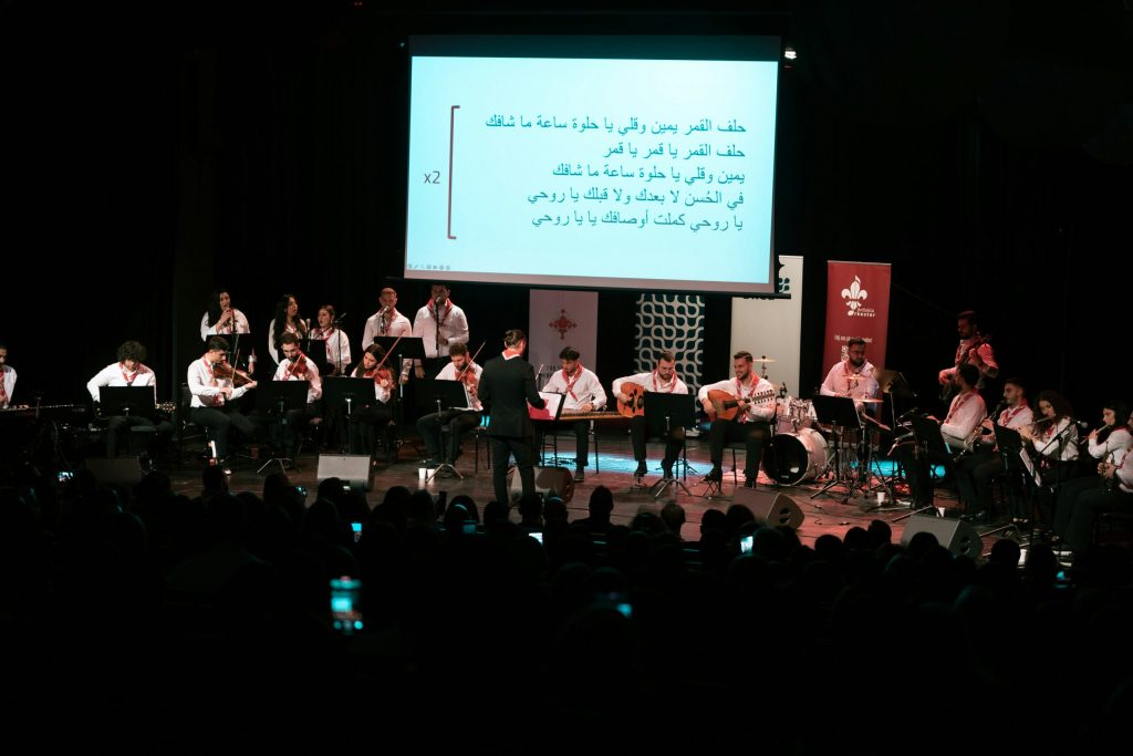 En stor musikensemble uppträder på scenen med texter projicerade bakom sig på arabiska.