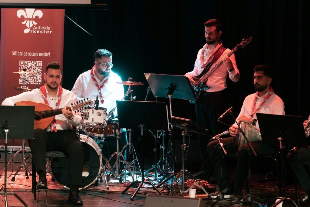 Fem musiker står på scenen och spelar olika instrument, med notställ och en röd orkesterfana bakom sig.